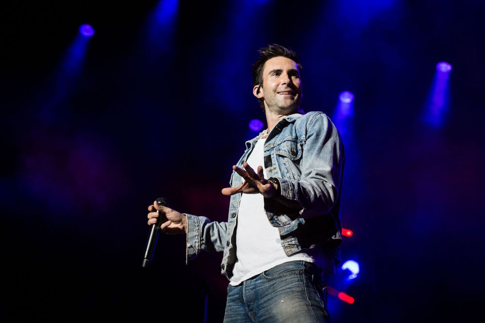 A performer holds a microphone on stage wearing a light denim jacket under dramatic blue stage lighting. Maroon 5. Adam Levine