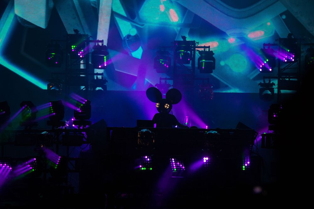 A dark concert venue illuminated with vibrant purple and blue lights while silhouetted audience members watch the stage. Deadmau5
