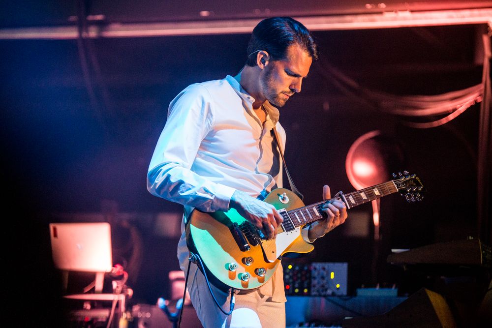 Musician in white shirt performs on stage with electric guitar under dramatic blue and red lighting.
