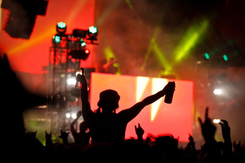 A silhouetted concert crowd member holds up a phone with colorful stage lighting and smoke effects in the background.