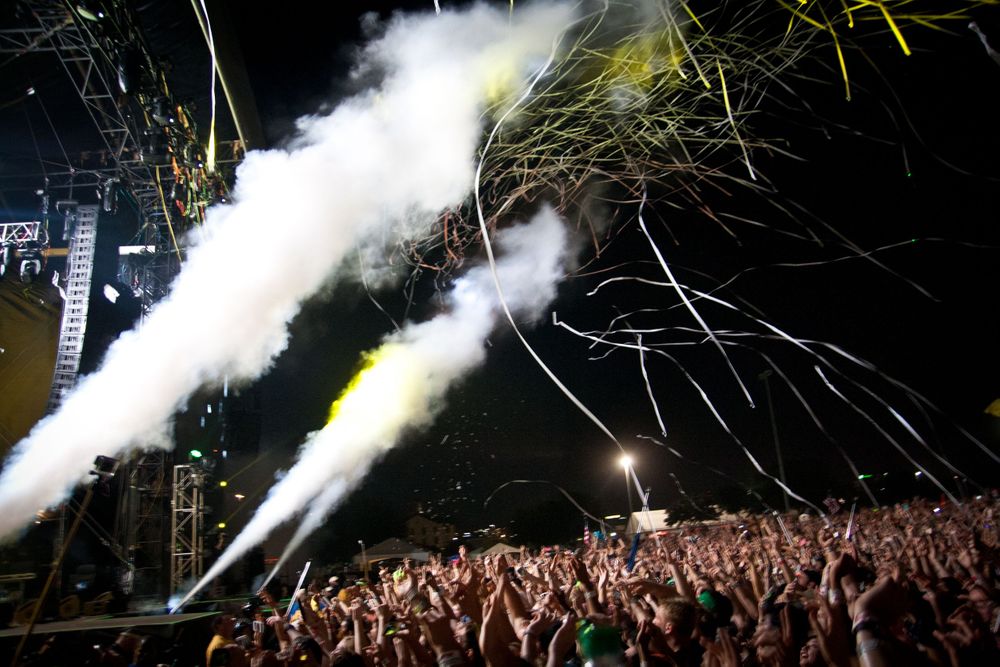 Concert crowd with raised hands under dramatic smoke machines and lighting effects at a music festival.