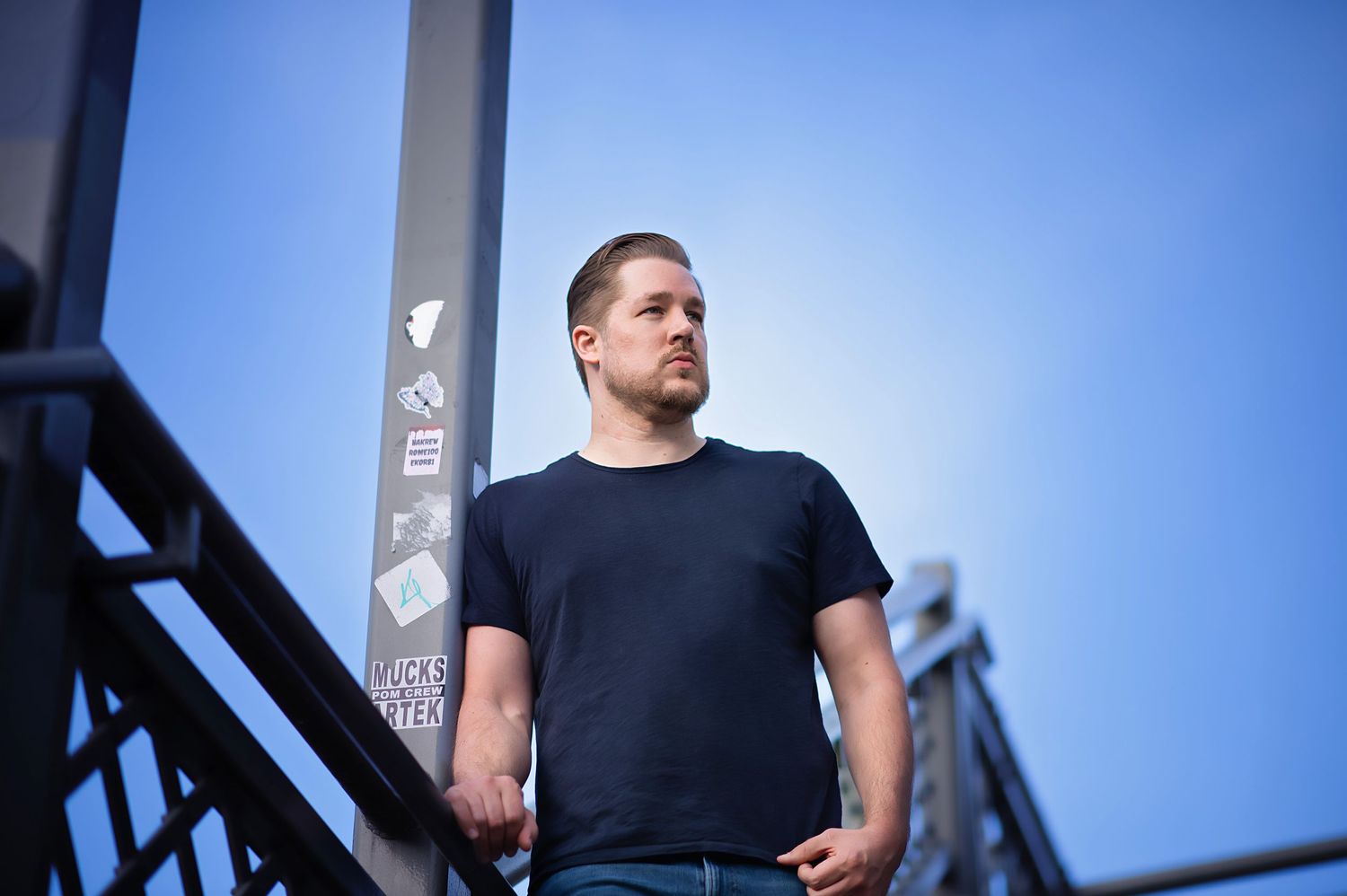 Person in black t-shirt stands against blue sky with industrial structure in background.