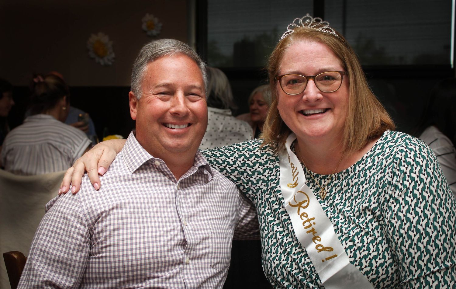 Two people pose together at an indoor celebration with one wearing a tiara and sash.