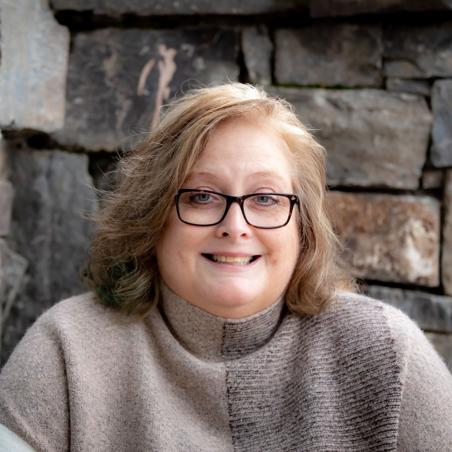 Portrait against stone wall wearing glasses and grey turtleneck sweater while smiling warmly.