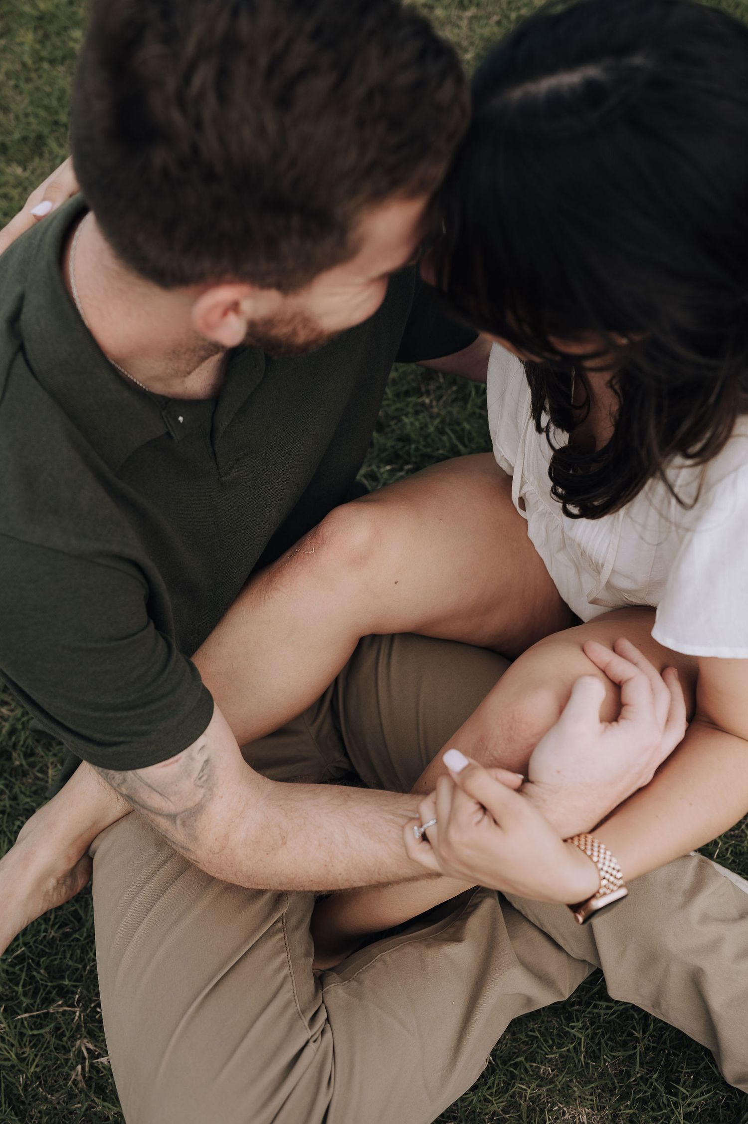 A couple sits close together on grass holding hands during an engagement session.