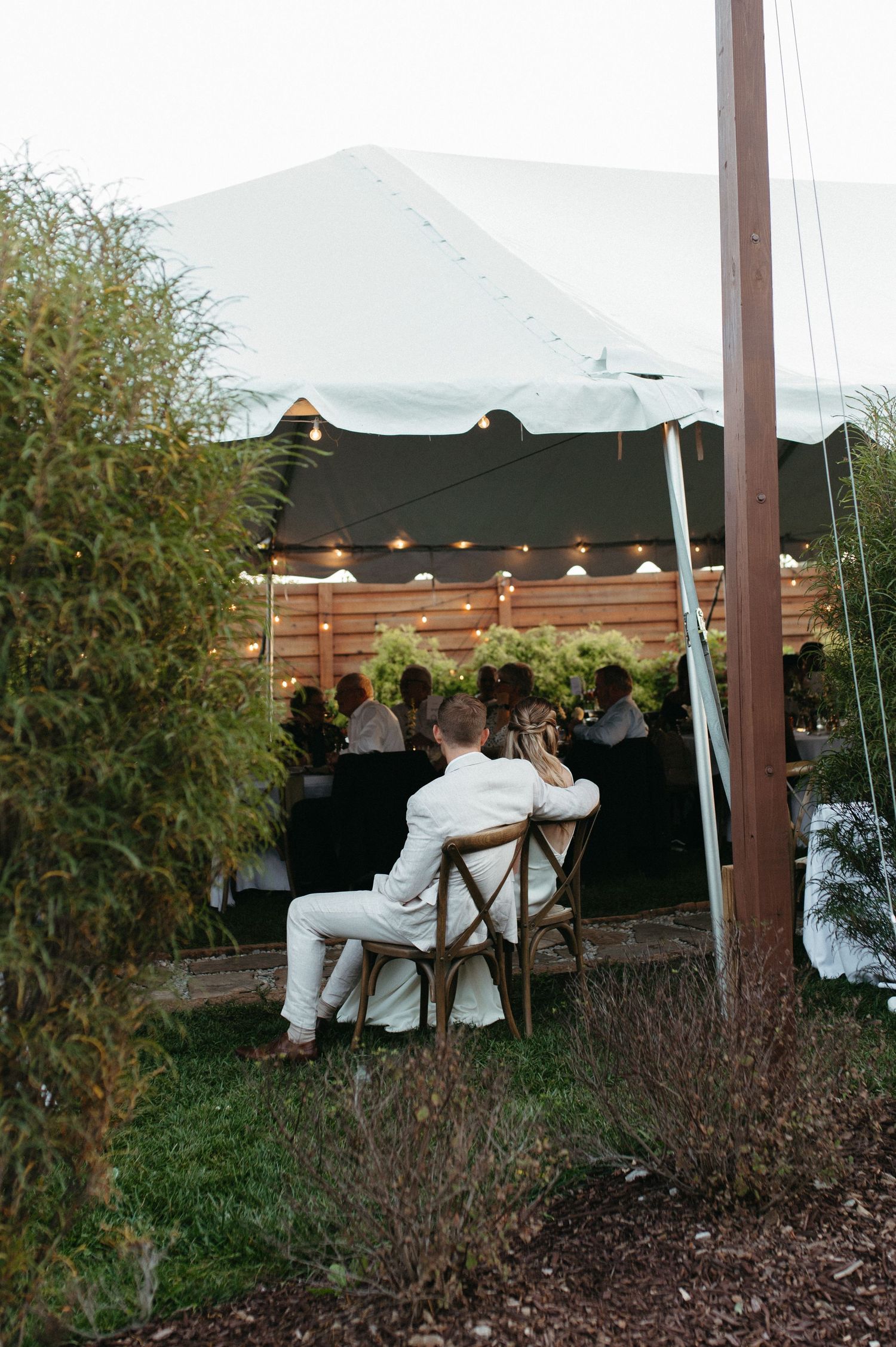 Wedding guests seated under a white tent with string lights at an outdoor reception.