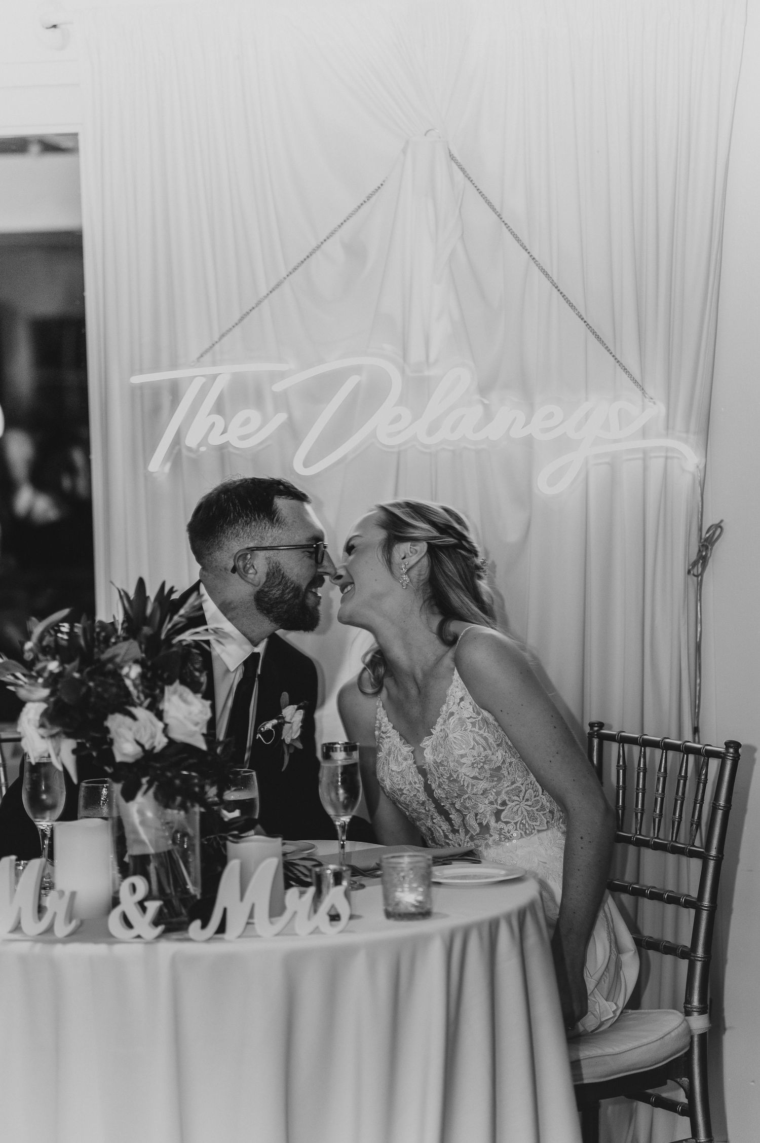 Black and white photo of newlyweds sharing an intimate moment at their reception table.
