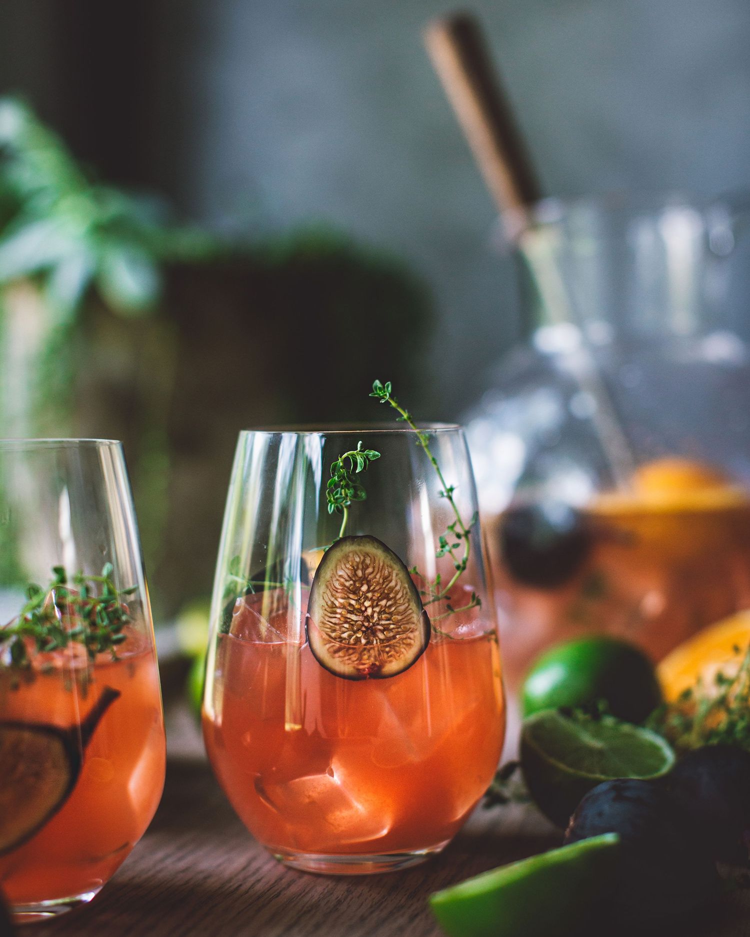 Three stemless glasses of pink fig and citrus cocktails garnished with fresh thyme on a rustic wooden surface.