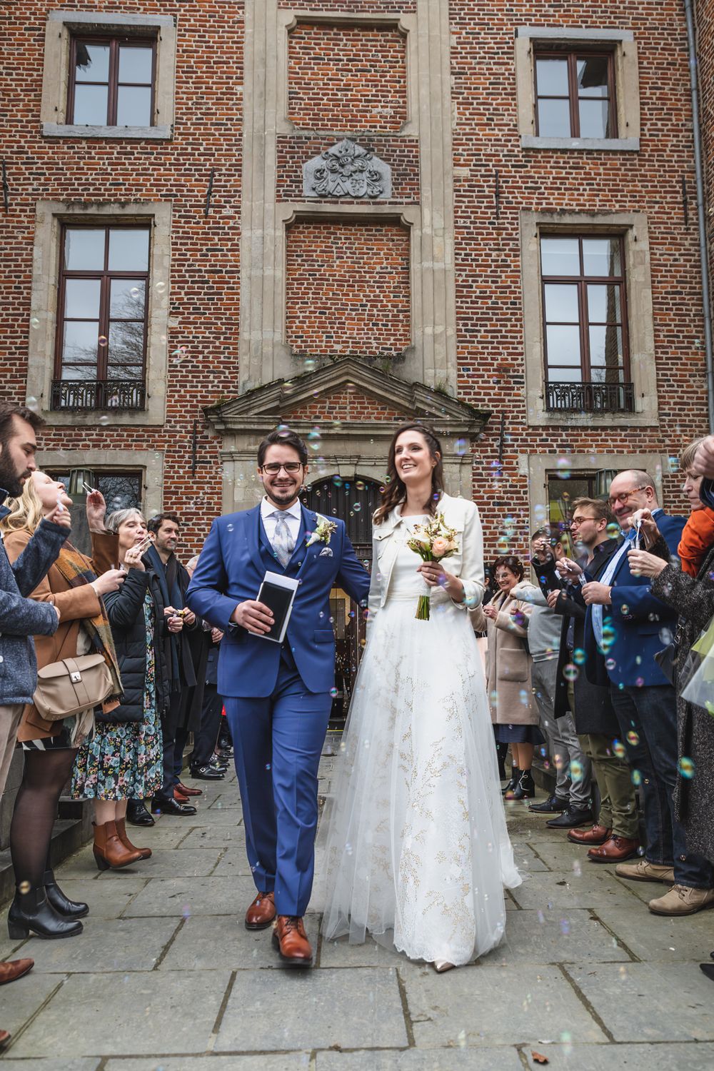 A newlywed couple exits a historic brick building as guests line the walkway.