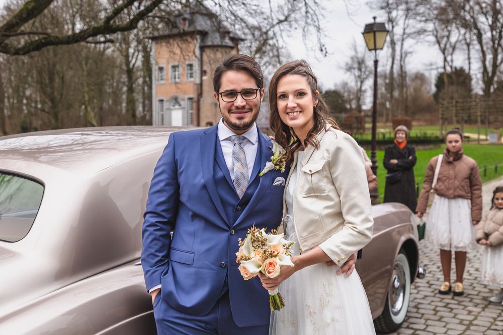 A wedding couple poses next to a vintage car outside a historic building.