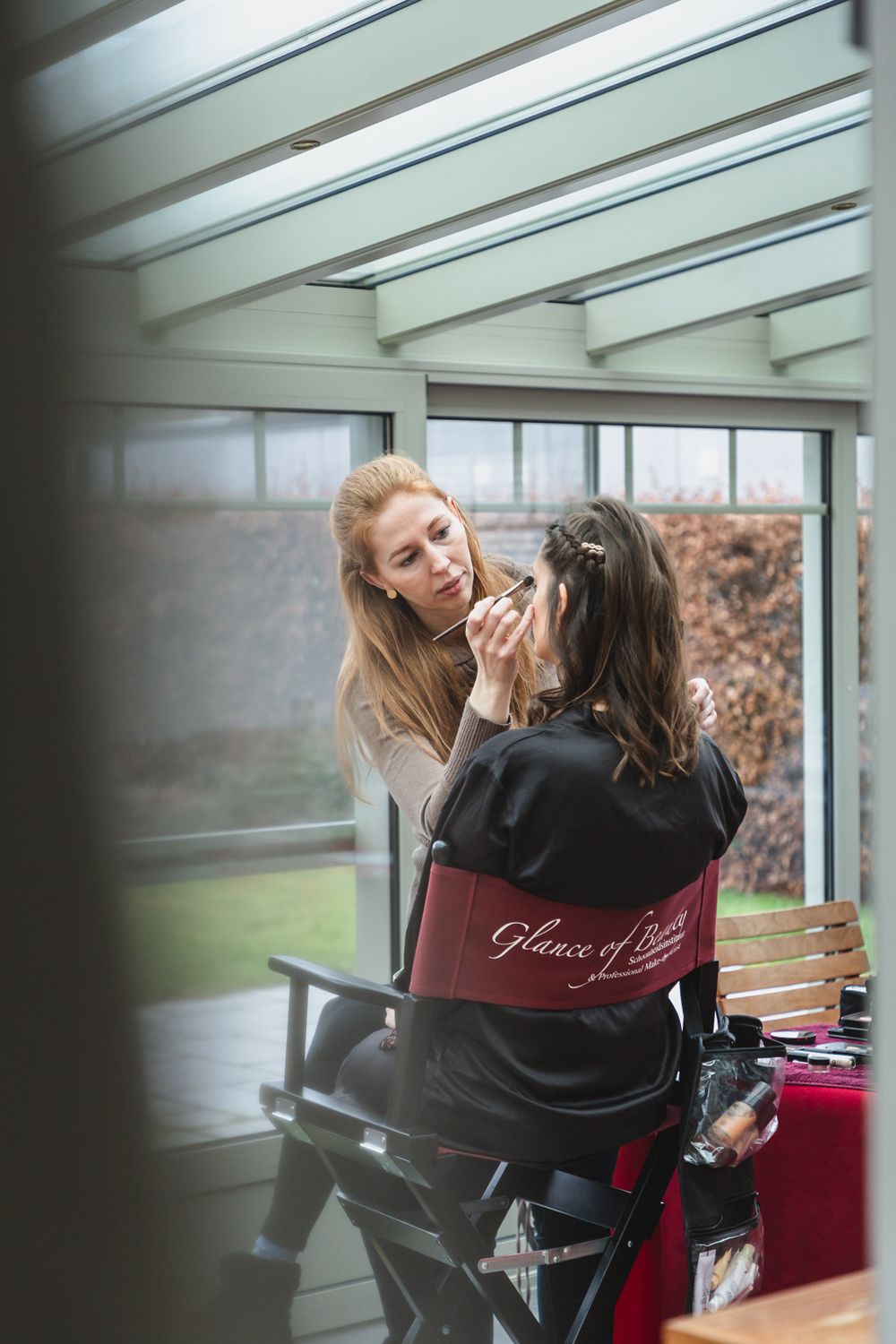 A makeup session takes place in a bright room with glass windows and beamed ceiling.