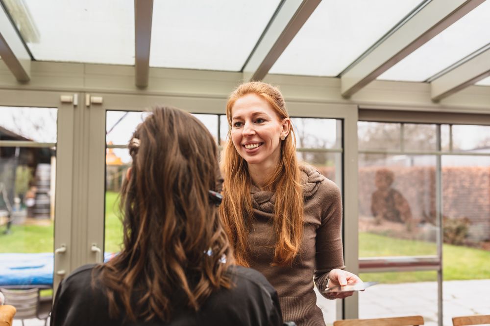 A makeup artist consults with a client in a sunlit conservatory space.