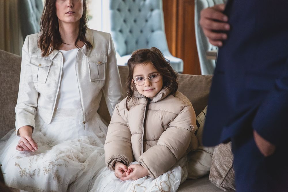 A child in a beige coat sits on a couch with adults in formal attire.