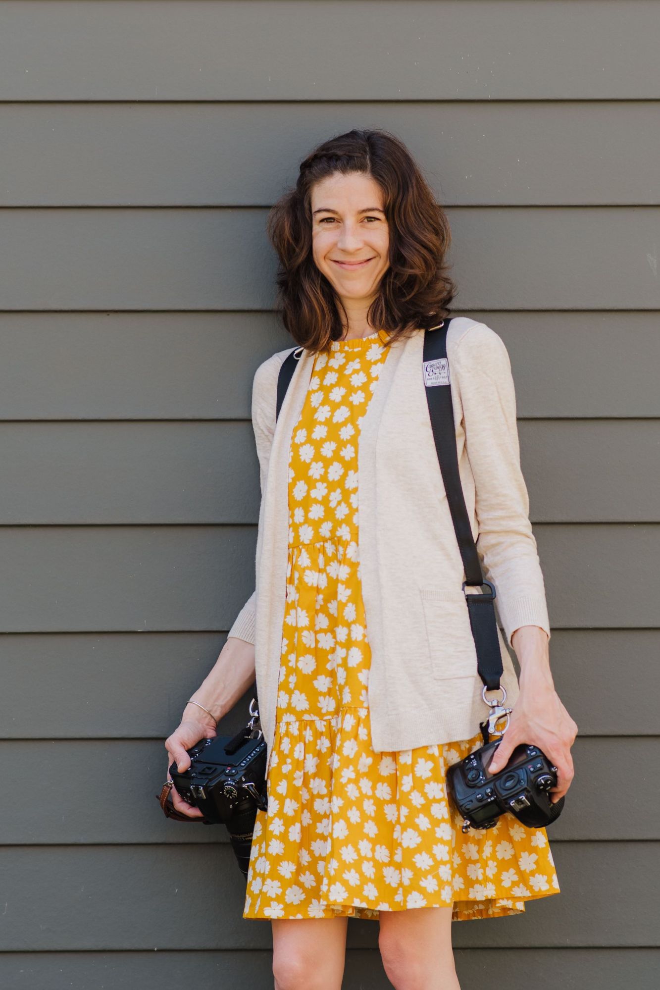 Photographer wearing a cheerful yellow polka dot dress with white cardigan and dual camera straps standing against gray wall.