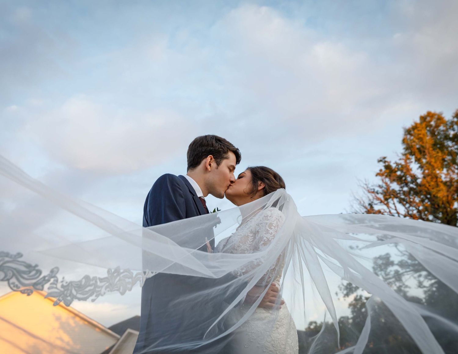 bride and groom kissing with veil sweeping in breeze on a fall autumn blue sky day