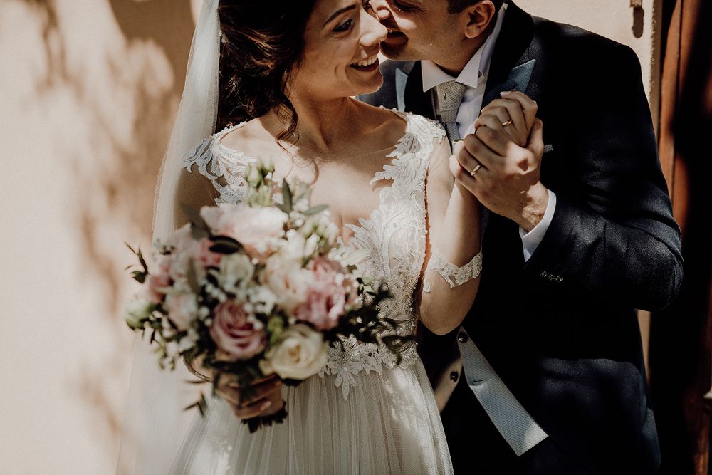 Smiling bride holding floral bouquet embraces groom in dark suit on wedding day.