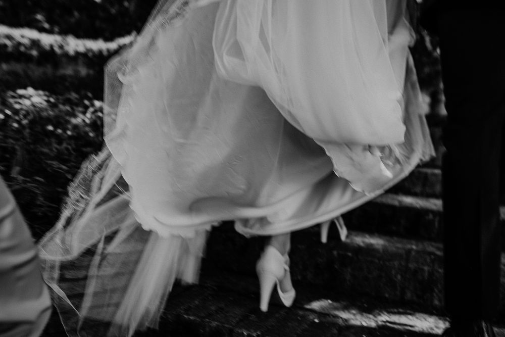 Black and white detail shot of bride lifting wedding dress while climbing stone steps.