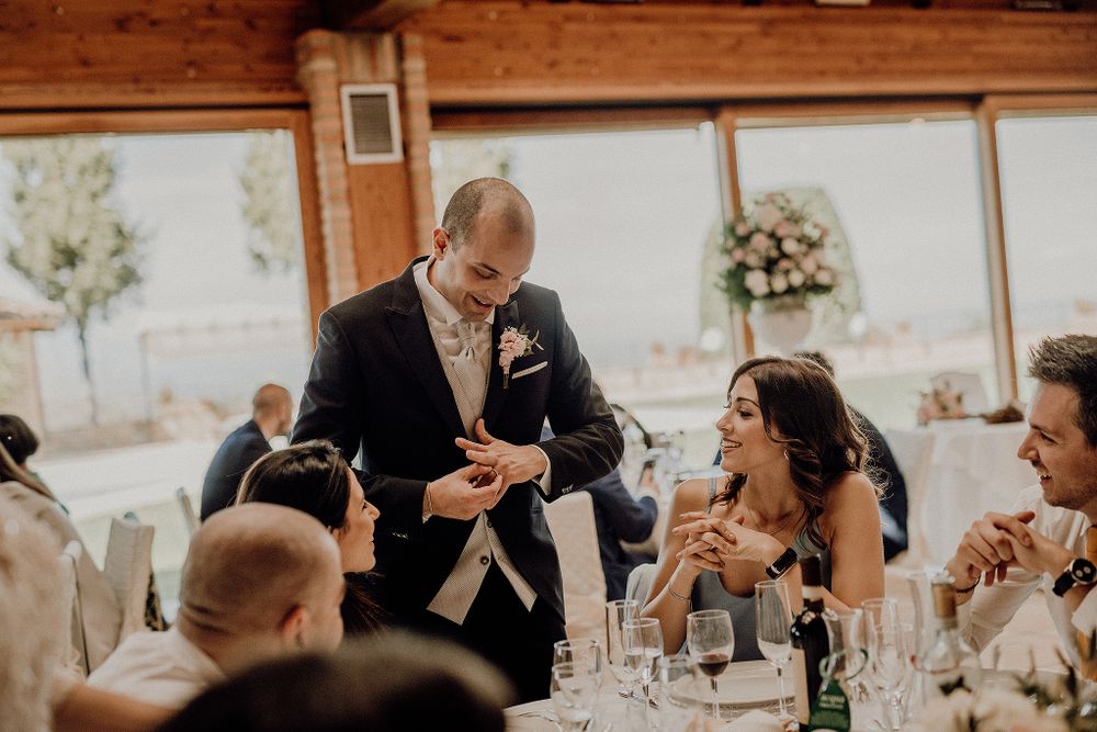 Groom in navy suit interacts with wedding guests at elegantly set reception tables with floral centerpieces.