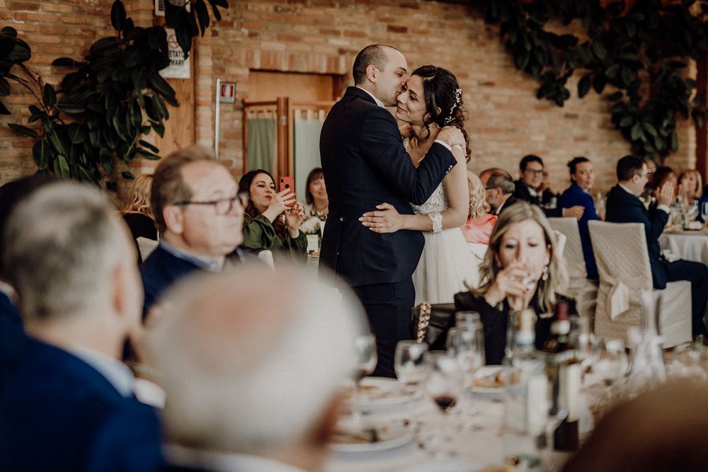 Newlyweds share a romantic first dance while wedding guests watch and applaud in a rustic reception hall.