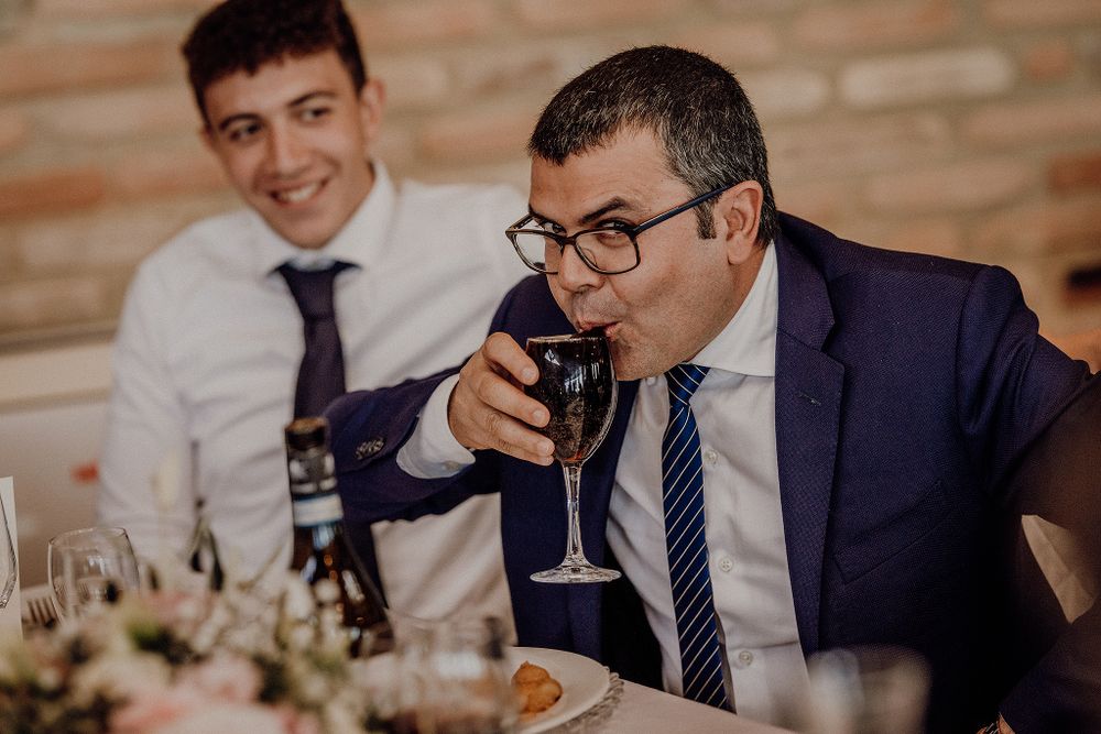 Wedding guest in navy suit enjoys a glass of red wine at a festive reception dinner table.