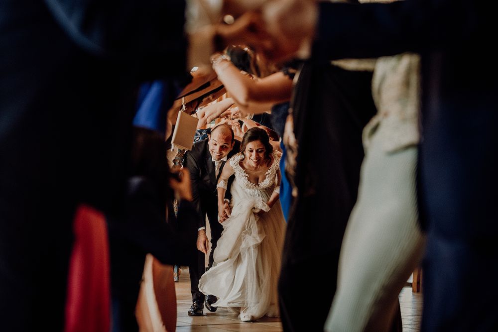 Bride in white gown walks through a celebratory tunnel of raised hands formed by wedding guests.