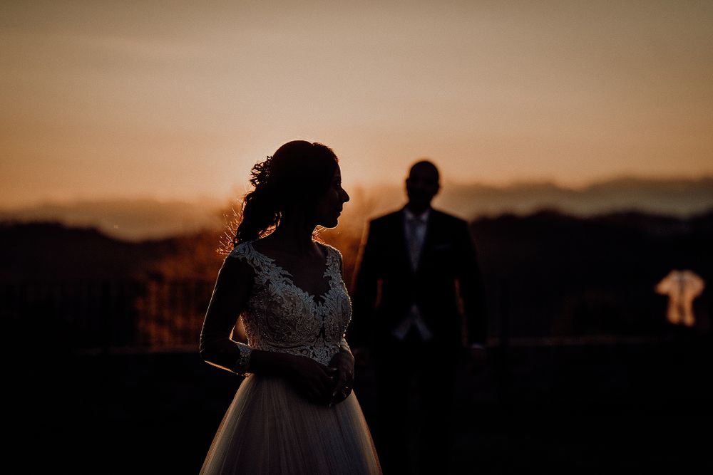 Bride in white gown silhouetted against golden sunset with groom standing in background.