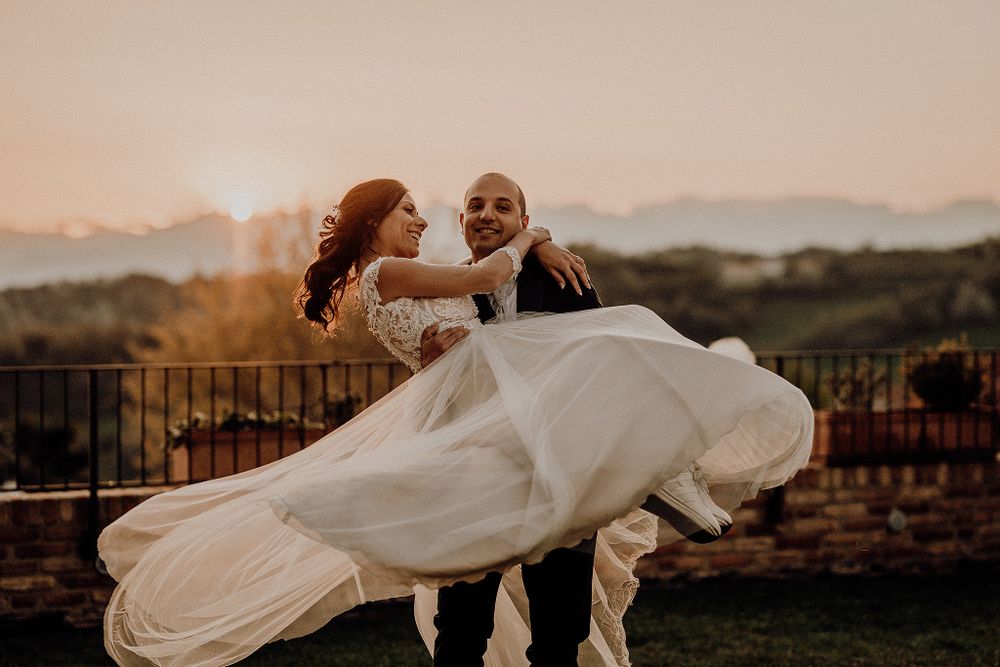 Groom lifting bride in elegant white gown at sunset with scenic landscape behind.