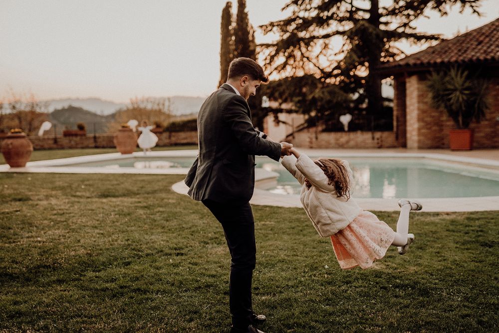 Father spinning young girl in colorful dress near poolside garden at dusk.