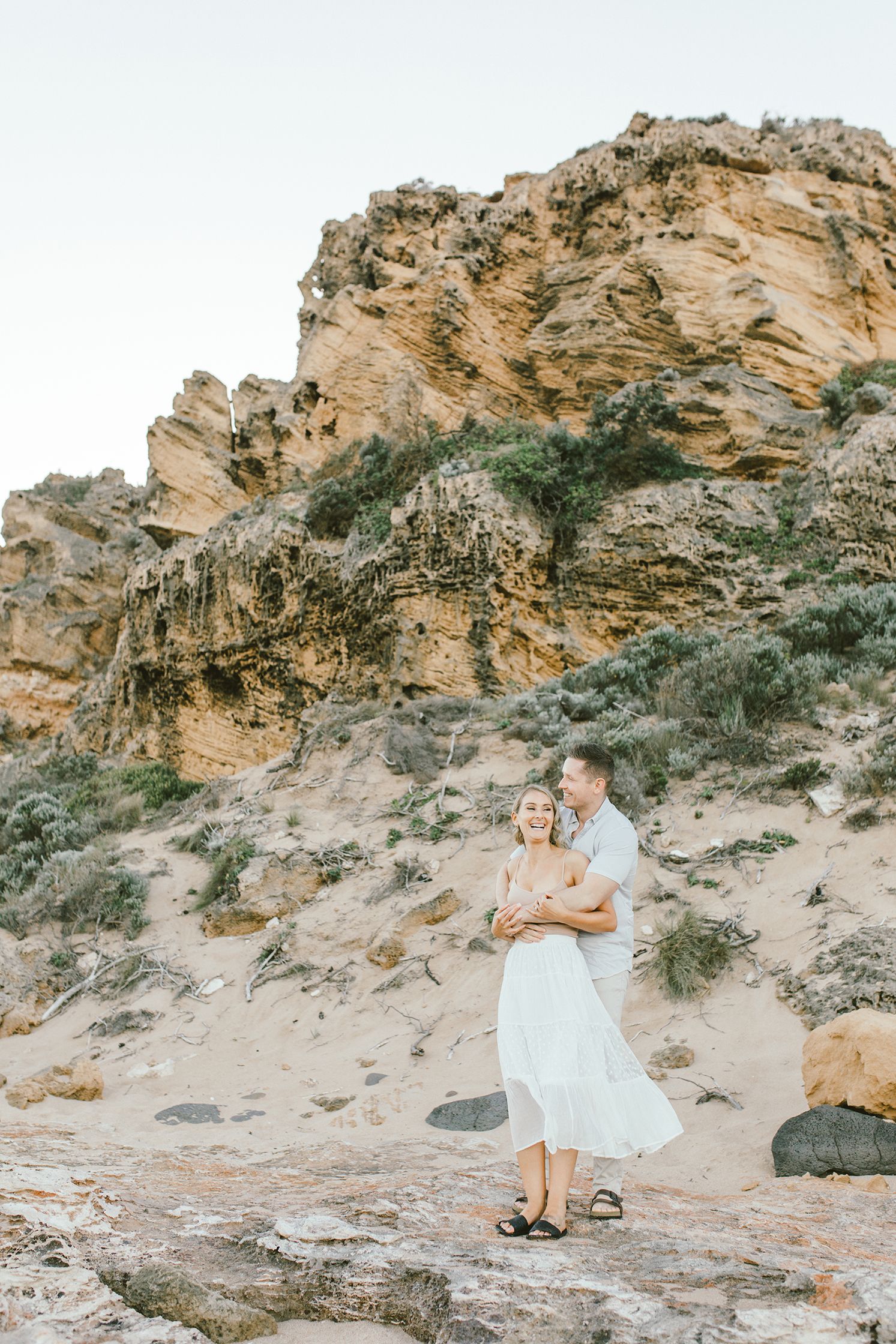 A couple poses against dramatic coastal cliffs during a beach photoshoot at sunset.
