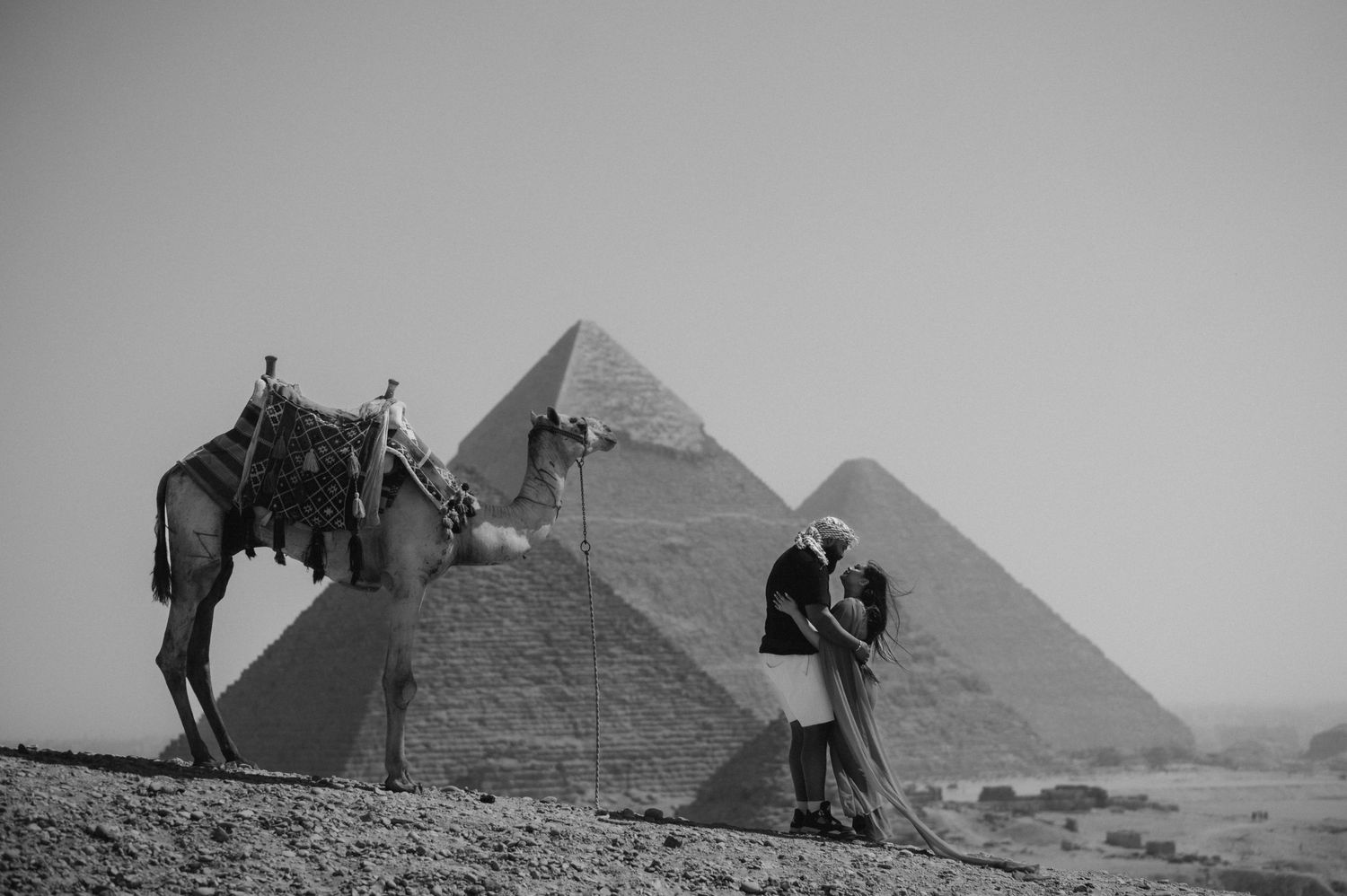 Romantic couple poses near a camel with the Egyptian pyramids in the background.