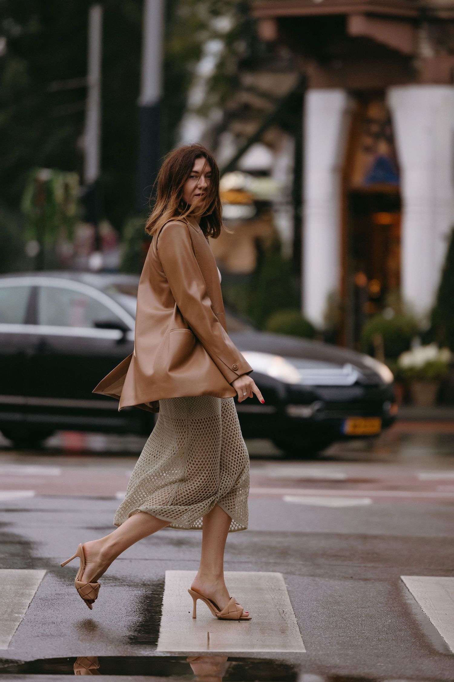 Stylish figure in camel blazer and beige knit skirt walking across a wet city street.