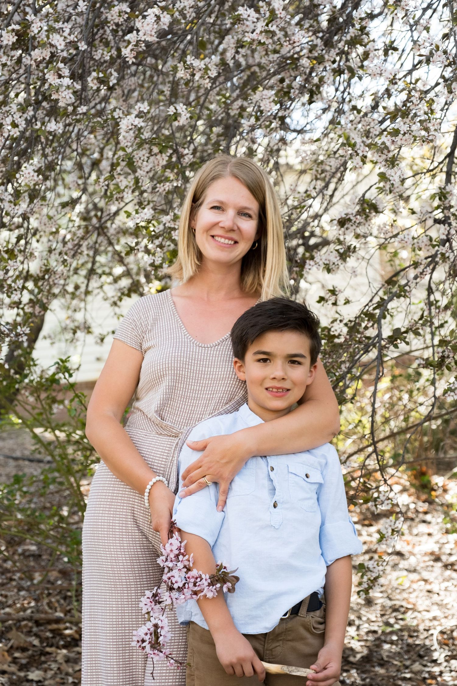 A parent and child pose together outdoors against a flowering tree backdrop on a sunny spring day.