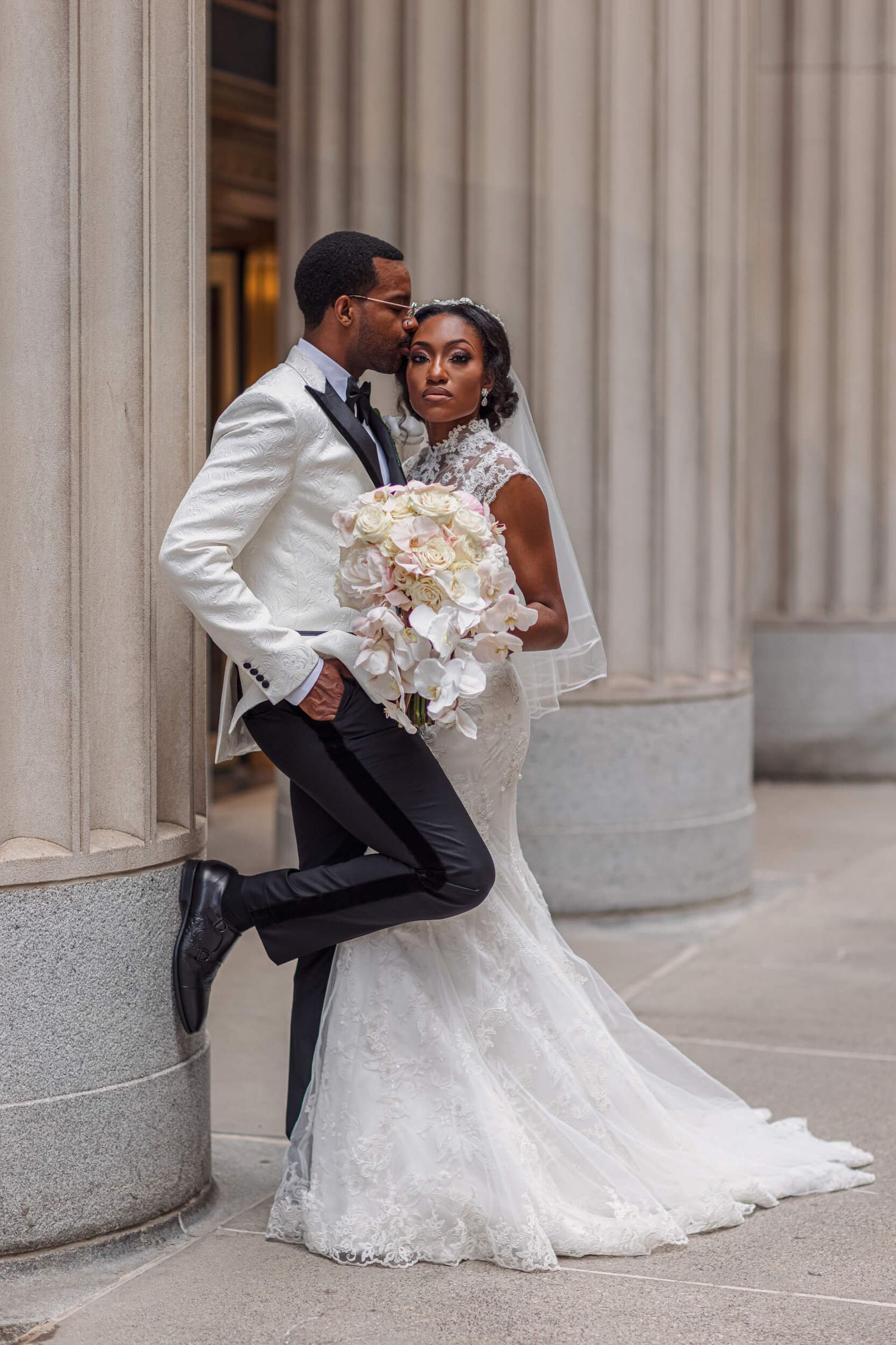 A bride in a white lace gown and groom in white tuxedo pose elegantly against classic stone columns.