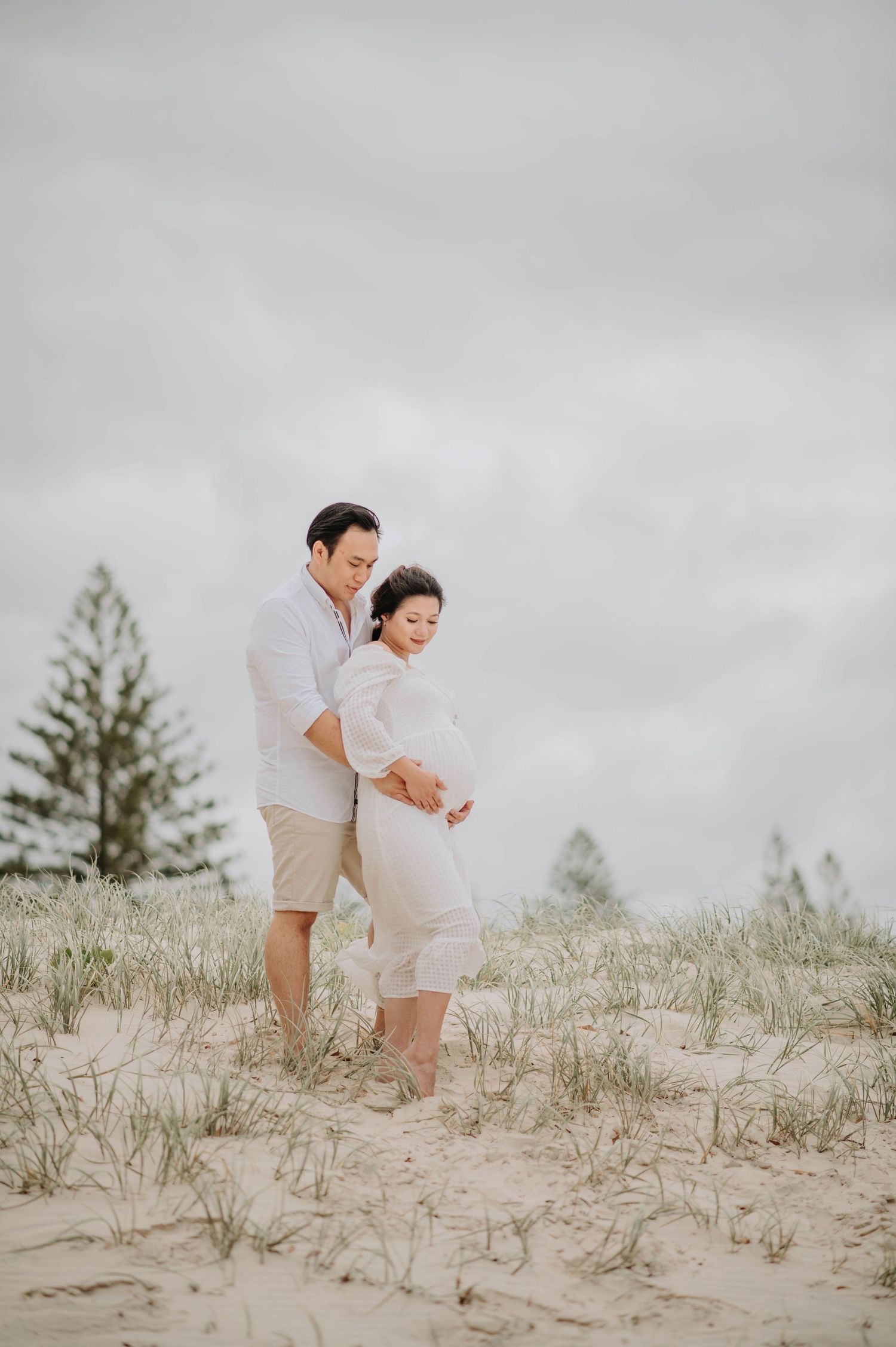 A couple embraces on sandy dunes with pine trees and overcast sky in the background.
