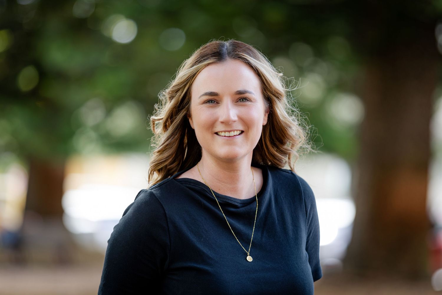 Outdoor portrait of someone in a navy top with a necklace against a natural background.