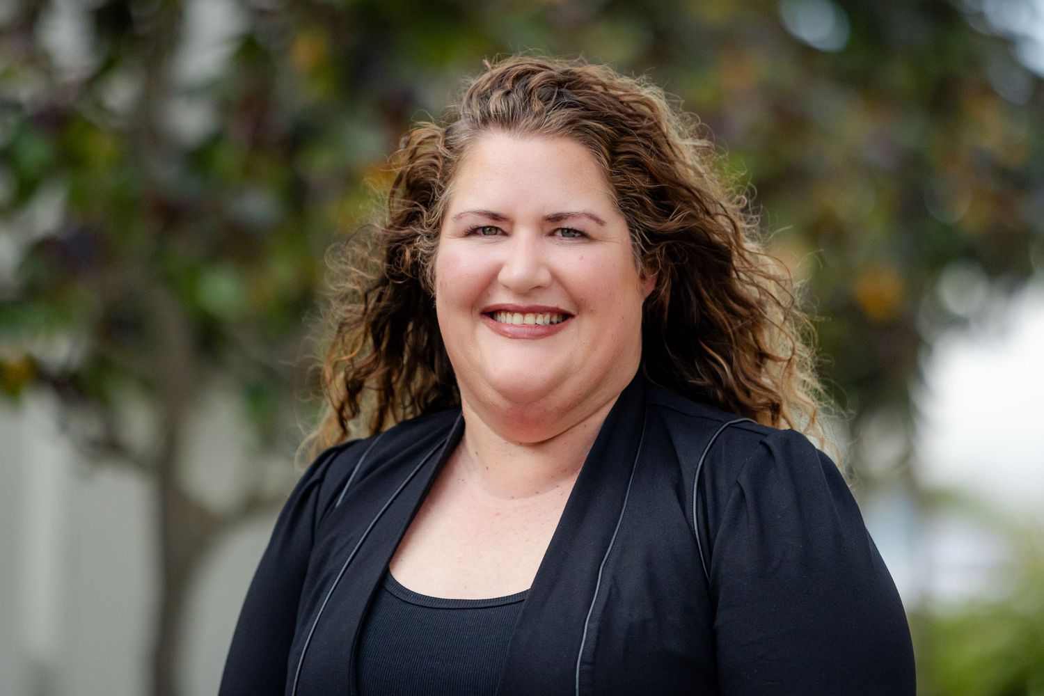 Professional headshot of someone wearing a black blazer against a blurred outdoor background.