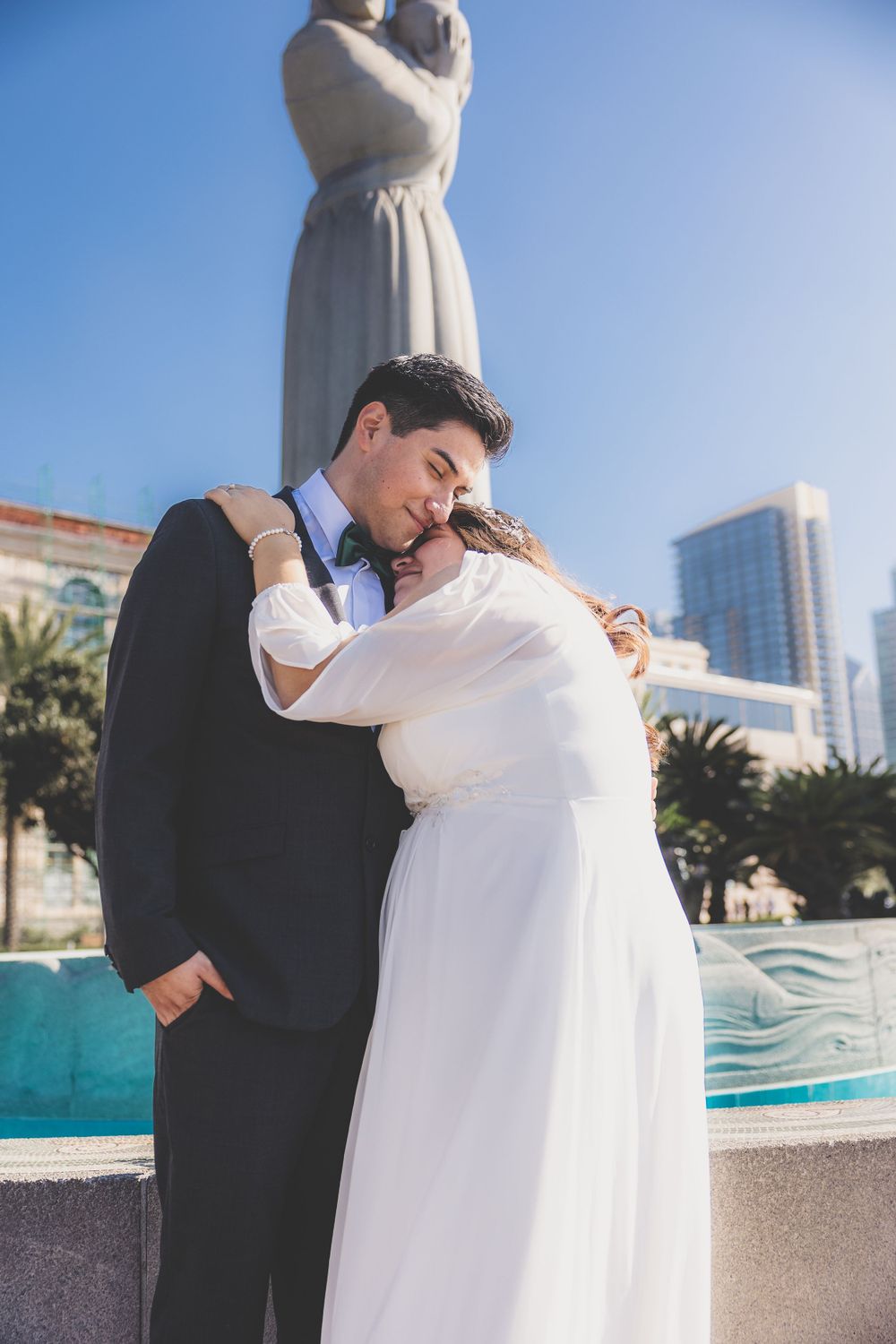 Bride and groom sharing tender embrace near decorative fountain statue under bright blue sky.