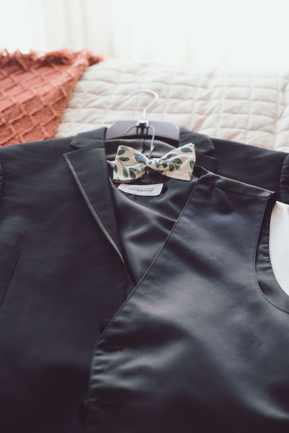 Black groom tuxedo jacket with floral bow tie on hanger displayed on bed.