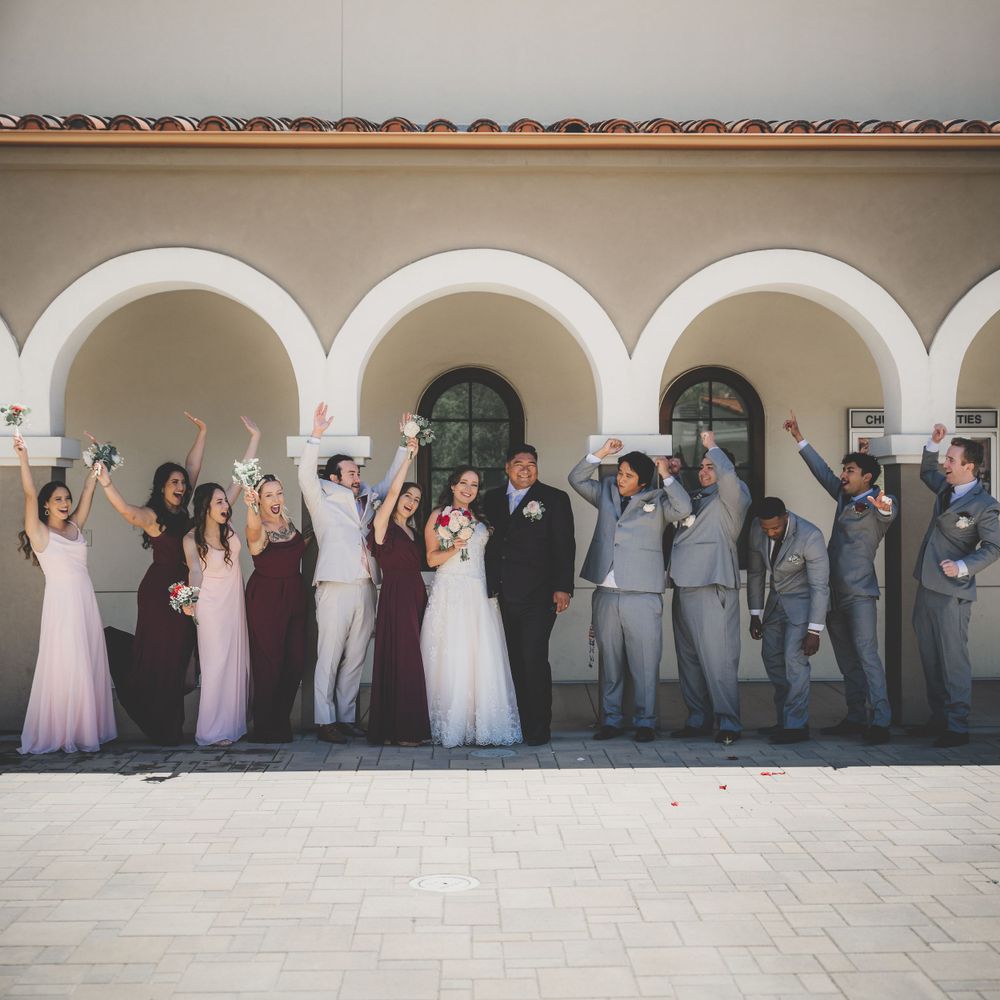 Wedding party celebrating under white arched colonnade with bridesmaids in burgundy and pink dresses.