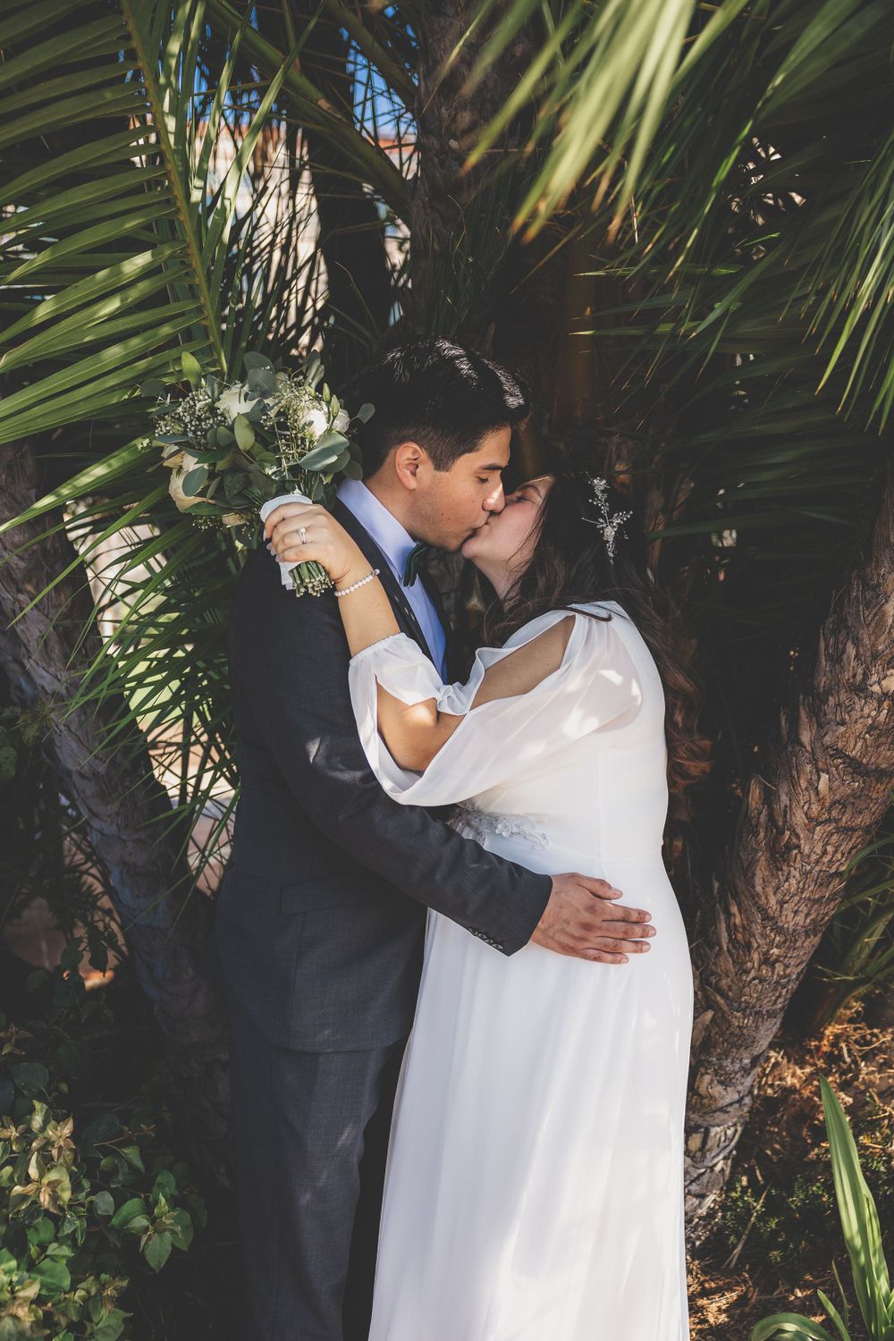 Bride and groom sharing romantic kiss beneath lush tropical palm tree fronds outdoors.