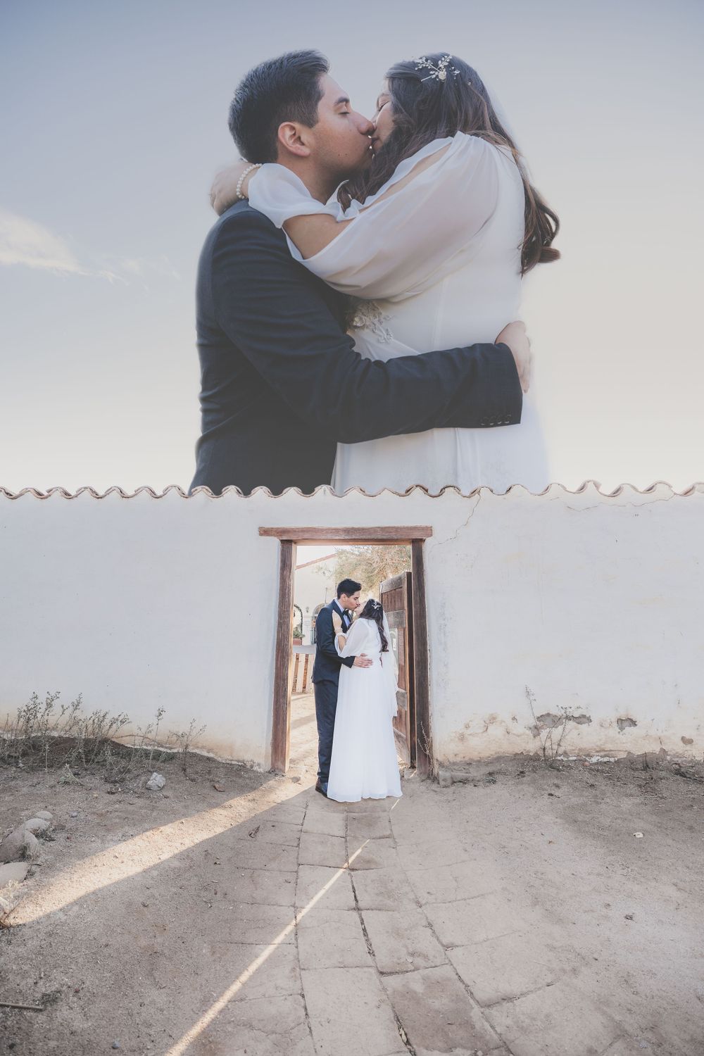 Double exposure composite of couple kissing above rustic white building doorway on wedding day.
