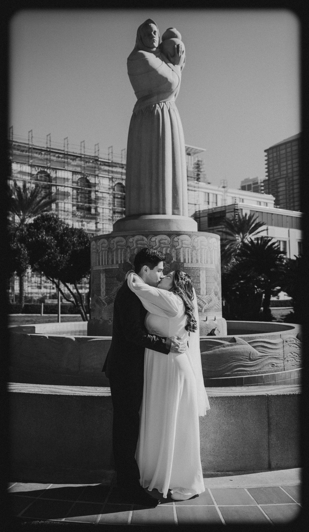 Black and white photo of newlyweds embracing before a classical fountain statue in urban plaza.