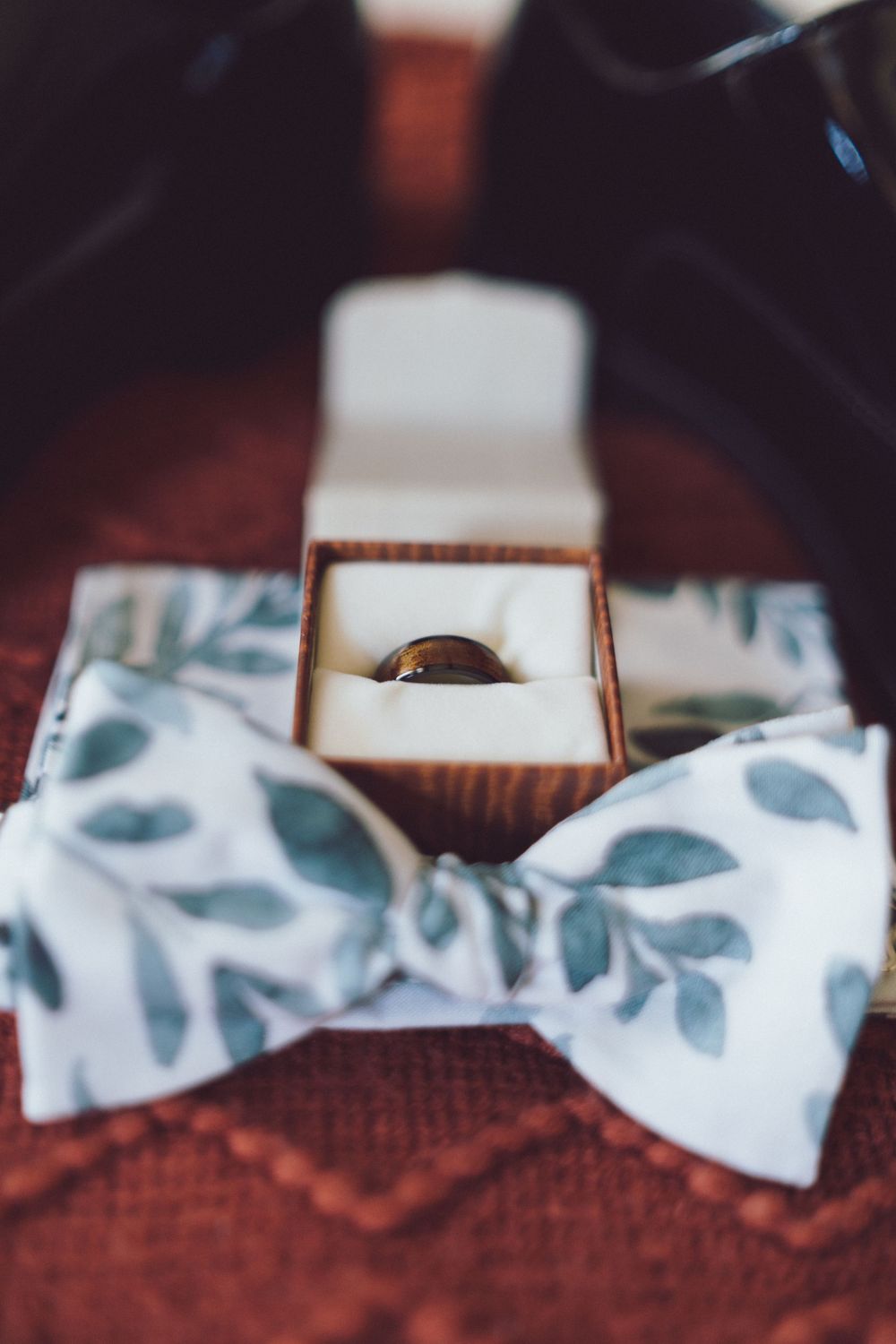 Close-up of groom wedding ring in box beside a white floral leaf patterned bow tie.