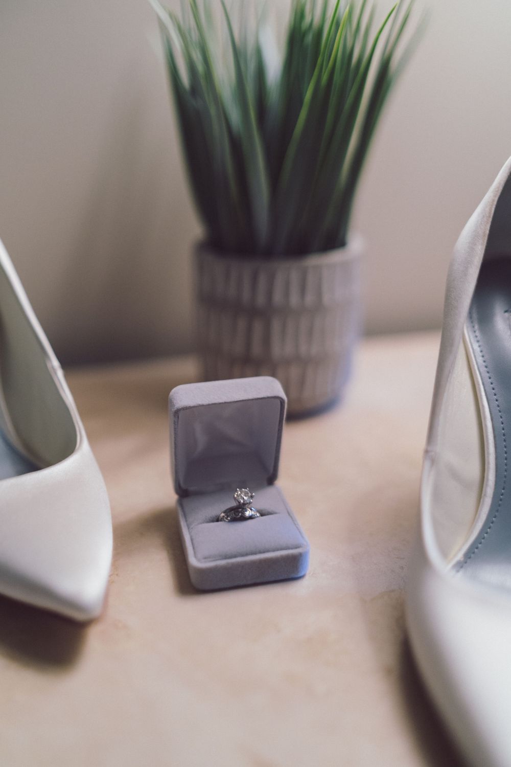 Diamond engagement ring in gray box displayed between white bridal heels and potted plant.