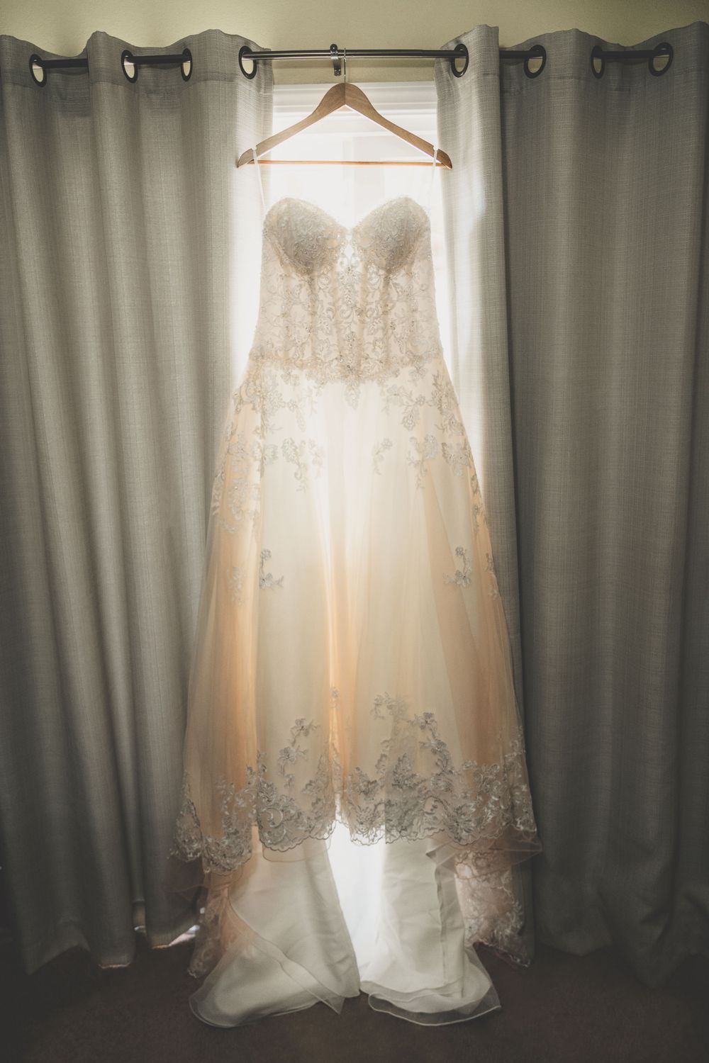 Elegant strapless lace and tulle wedding dress hanging in window light on wooden hanger.