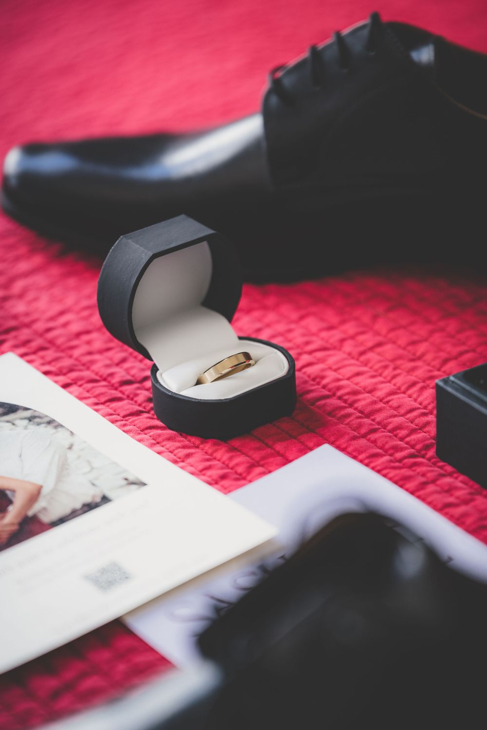 Gold wedding band in black ring box on red surface with black dress shoes and photos nearby.