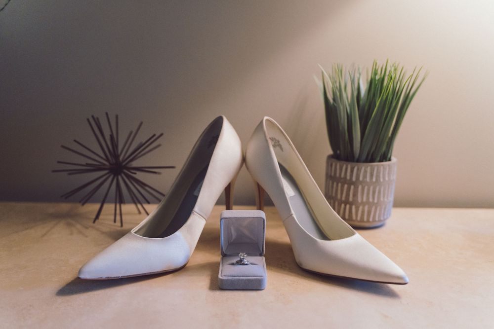 White bridal heels displayed with diamond engagement ring box and green potted plant on table.