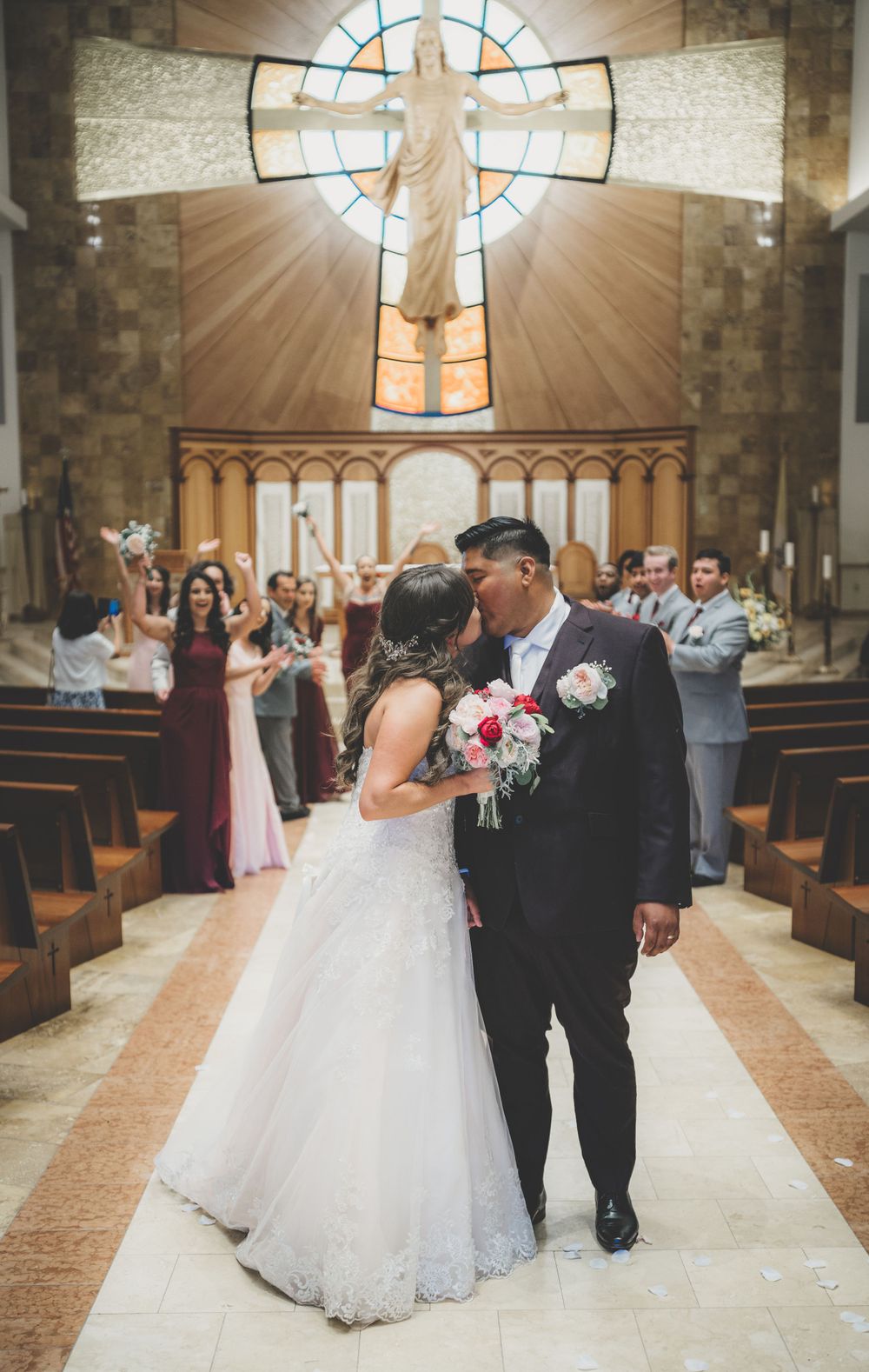 Newlyweds share first kiss in church aisle with stained glass cross and cheering guests behind them.