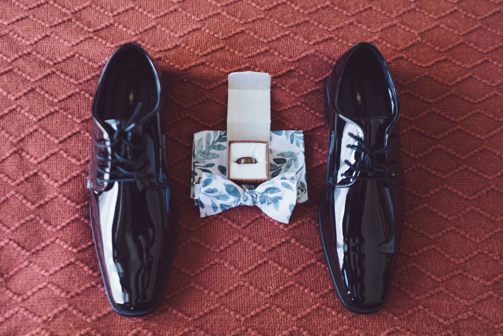 Groom wedding accessories flat lay with patent leather shoes, floral bow tie, and gold ring.
