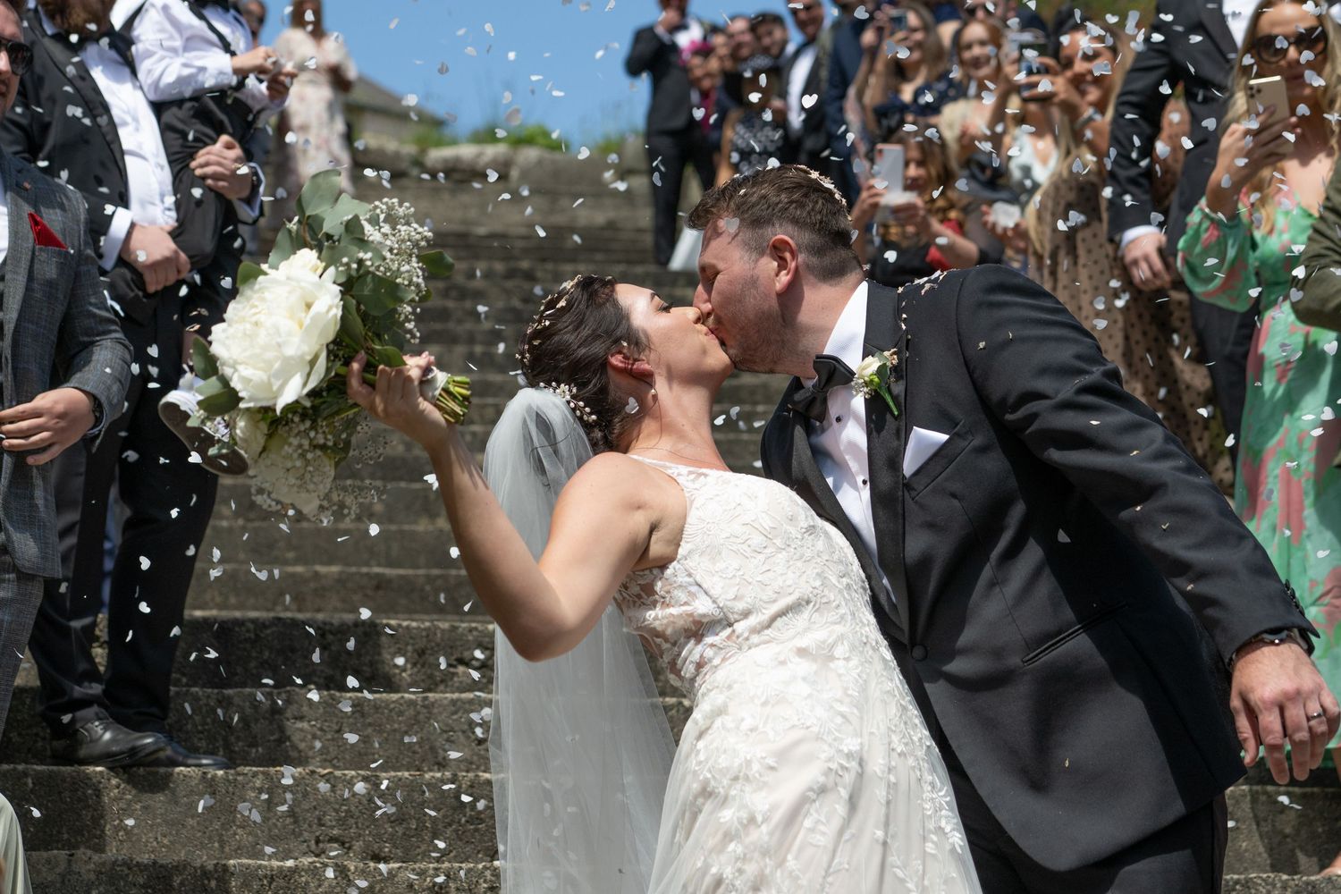 Newlyweds share a romantic kiss on stone steps while guests throw confetti.